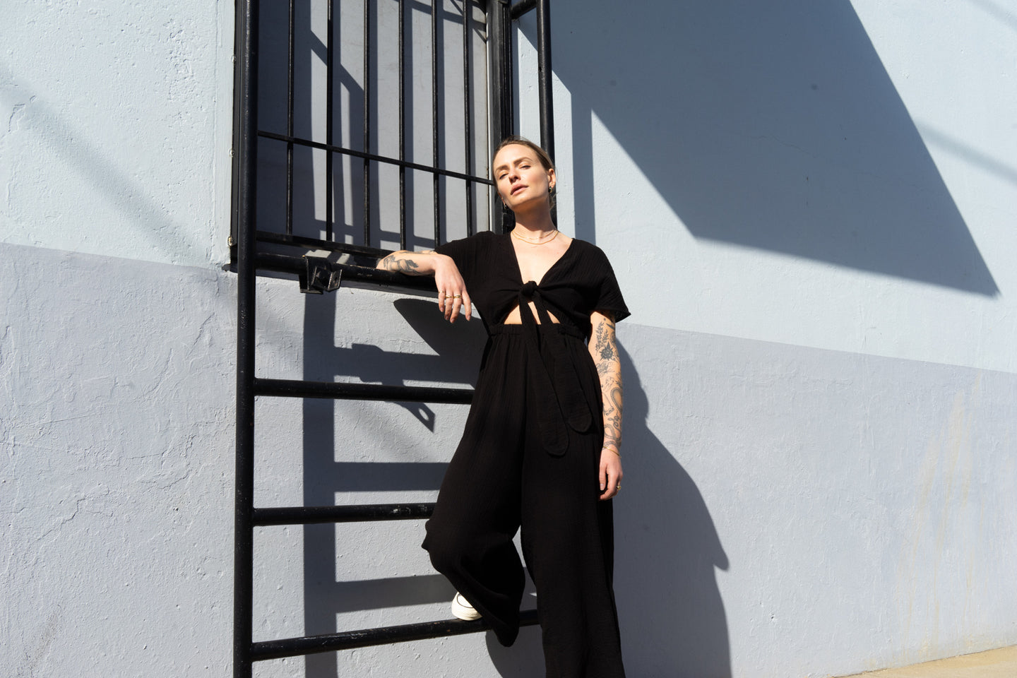 A woman in a black jumpsuit leaning against an industrial wall